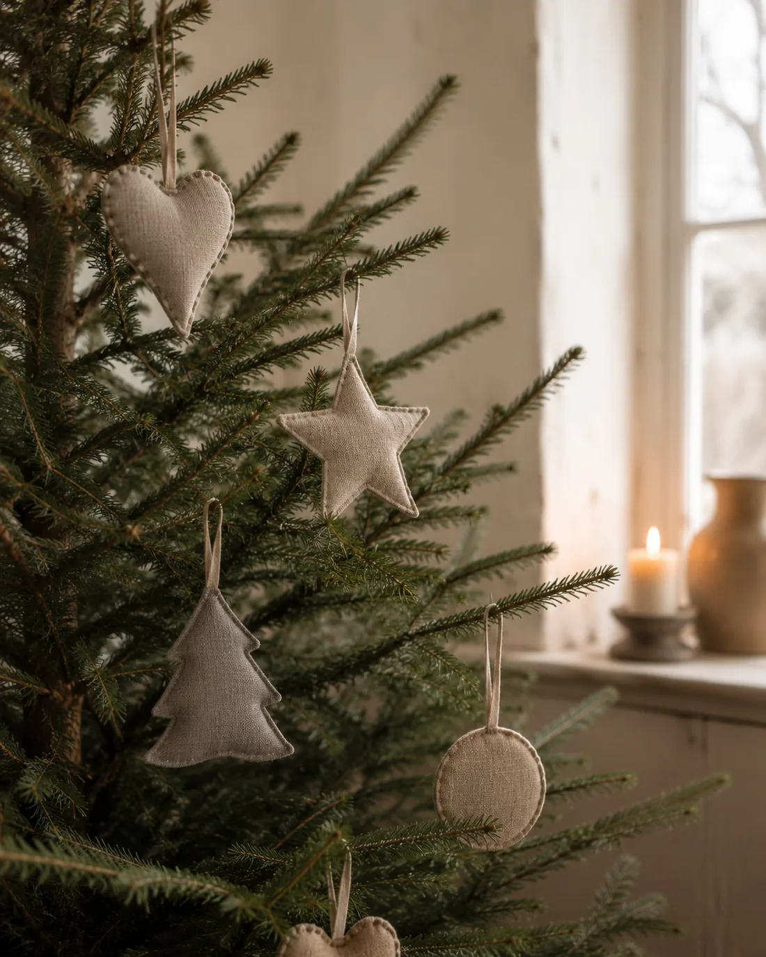 Vier flache Stoffanhänger in Naturleinen – Herz, Stern, Tanne und Kugel – weit verteilt an einem echten Tannenbaum, mit viel sichtbarem Zweig dazwischen